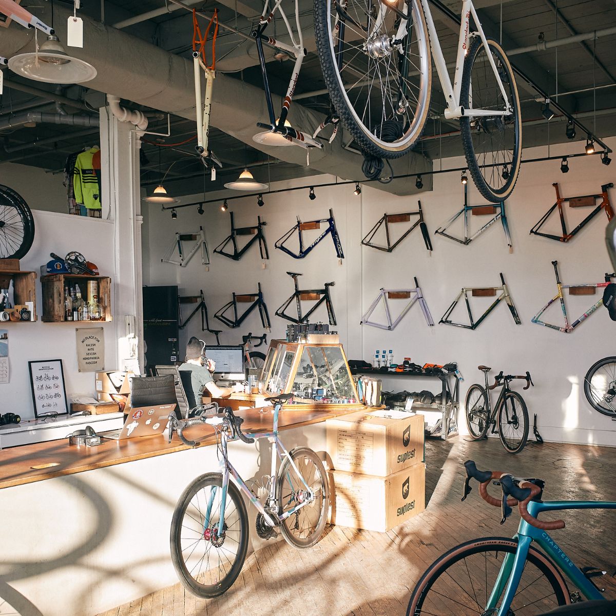 A bicycle shop with various bike frames displayed on the walls and complete bikes hanging from the ceiling. A counter with a laptop and cardboard boxes is in the middle, with sunlight streaming in through the windows.