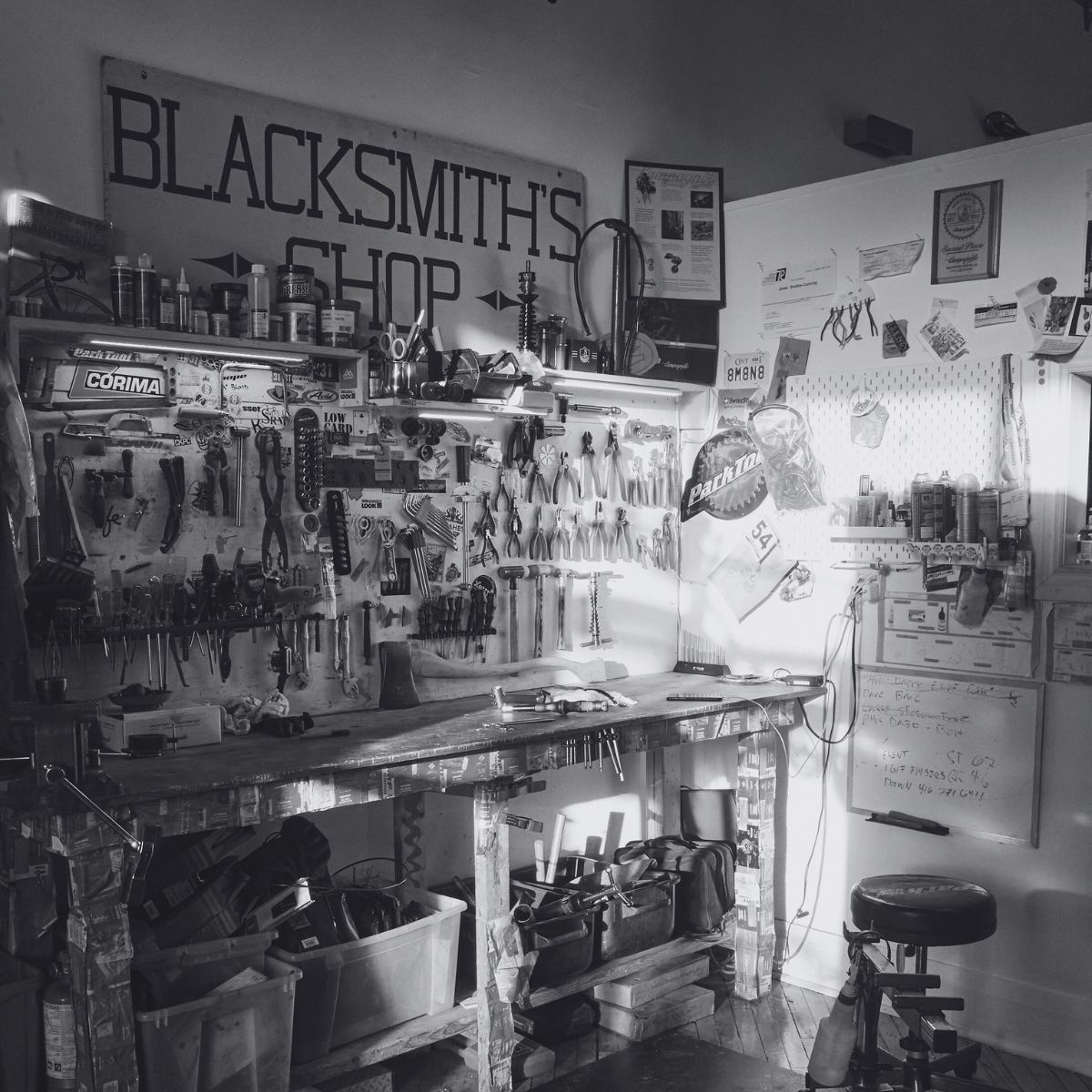 Black and white image of a cluttered workshop labeled "Blacksmith's Shop." The workbench is filled with various tools, shelves hold parts and containers, and the walls are covered with posters and notes. A stool sits in front of the bench.