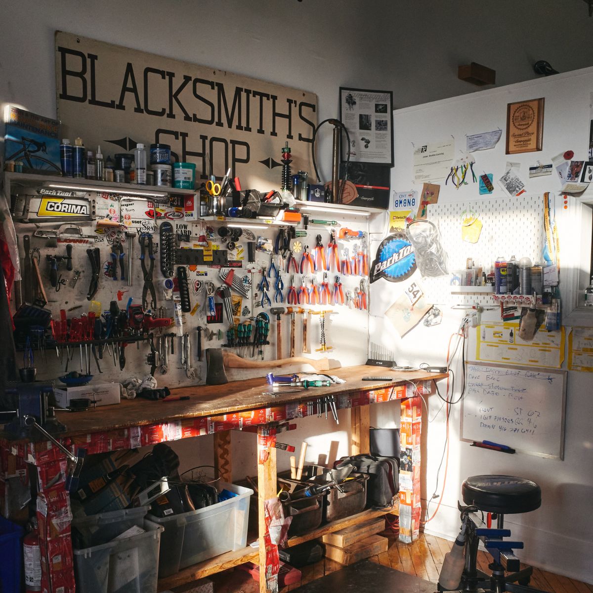 A sunlit workshop with a wooden workbench cluttered with tools, pliers, and various equipment. A "Blacksmith's Shop" sign hangs above. Walls display posters, notes, and a clock. A stool sits nearby, and sunlight streams through a window.