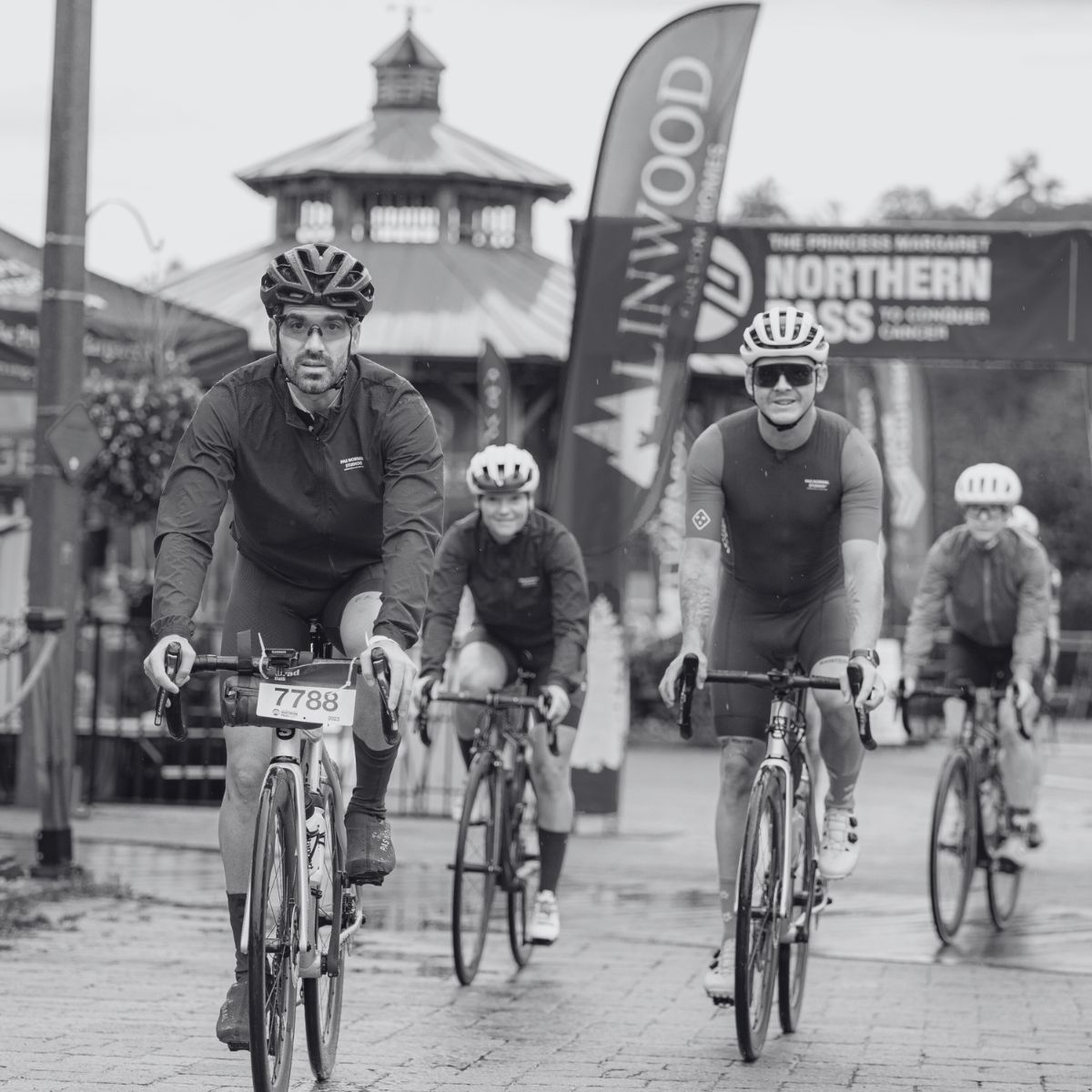 Cyclists participate in a race, riding along a paved street. Four riders are visible in the foreground, wearing helmets and numbered jerseys, with event banners and a gazebo in the background. The image is in black and white.