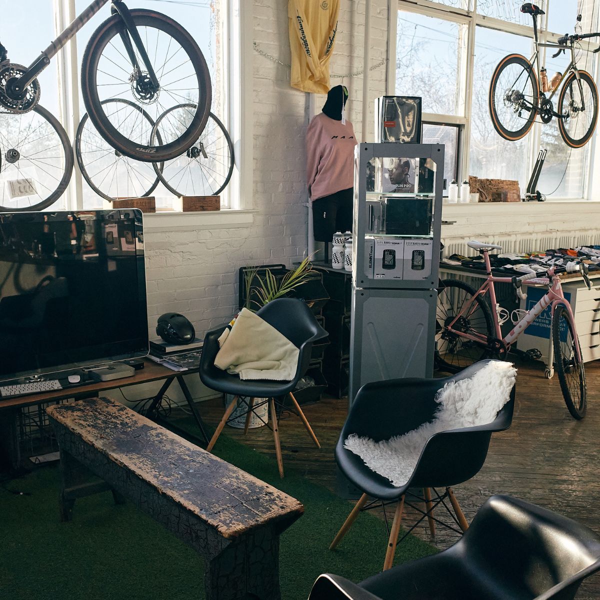 A cozy room featuring a rustic bench and black chairs with cushions around a small table. Bicycles are mounted on a white brick wall above, with large windows providing natural light. A computer and a tall cabinet are seen in the background.