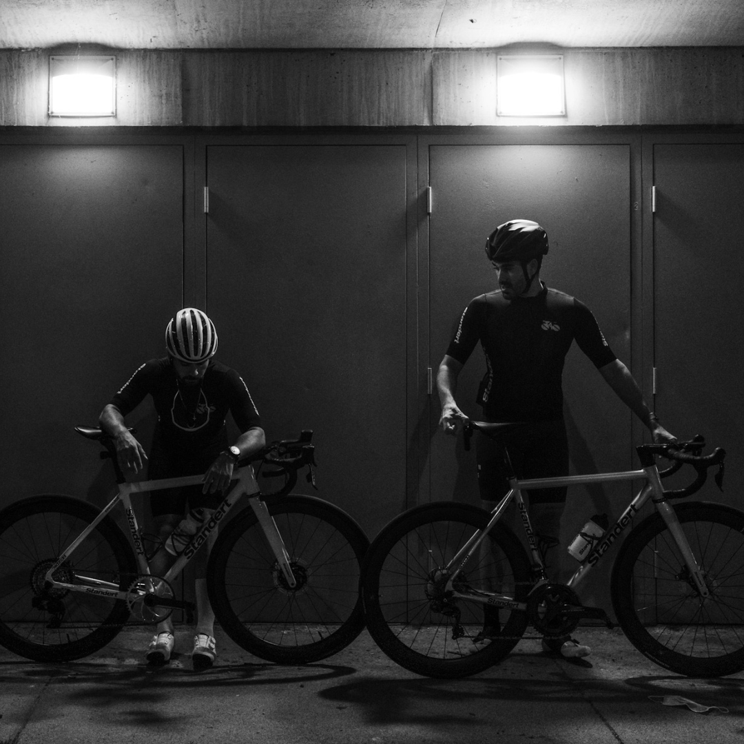 Two cyclists in helmets and cycling gear stand with their bicycles against a wall at night. The scene is lit by overhead lights, creating a dramatic contrast with the shadows.