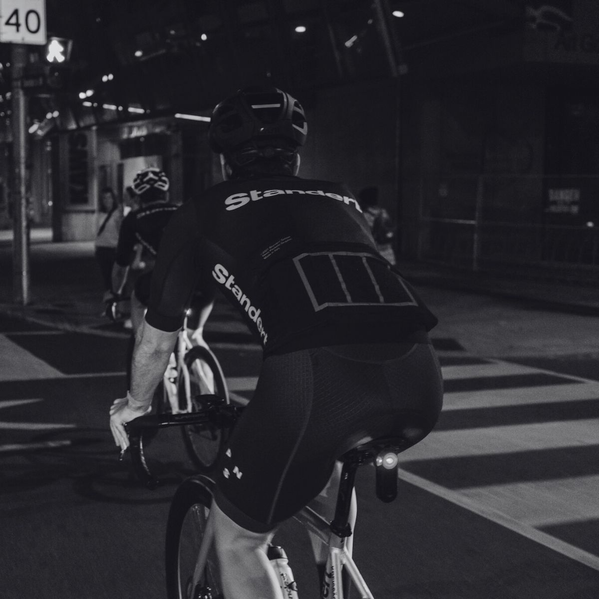 Black and white photo of cyclists riding on a city street at night. The cyclists wear helmets and jerseys with "Standert" printed on them. Street signs and illuminated buildings are visible in the background.