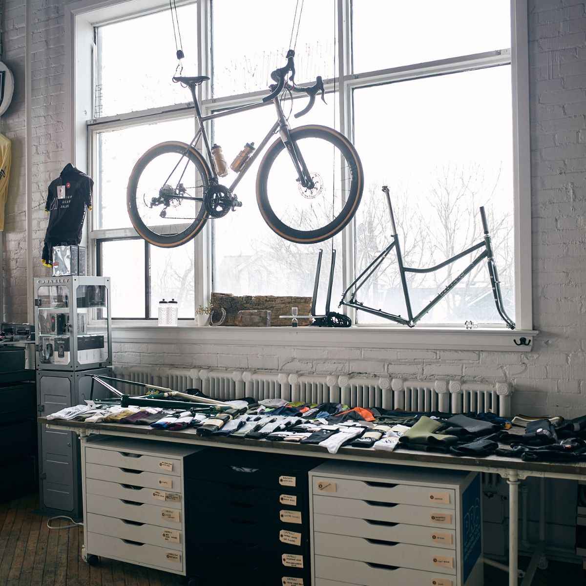 A bike hangs in front of large windows in a workshop. A bicycle frame is propped up beside it. A table underneath is covered with various tools and bike parts. Natural light illuminates the space.