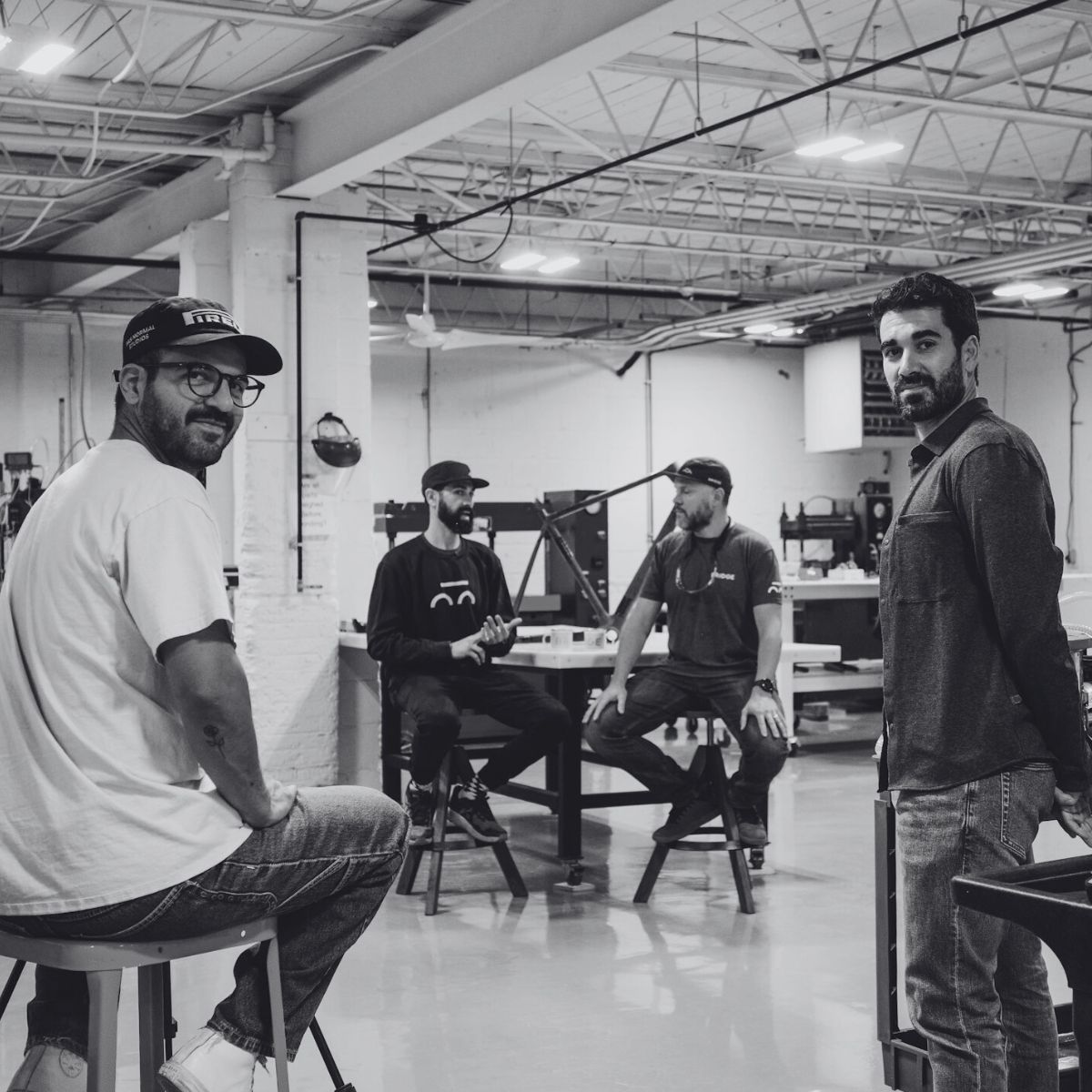 Four men sit and stand in a modern industrial workshop. They appear engaged in conversation. The space has high ceilings, exposed beams, and various tools in the background. The photo is in black and white.