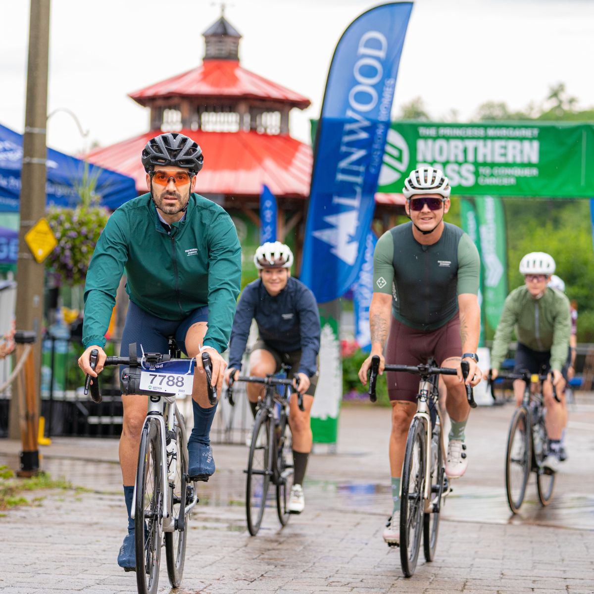 Cyclists in various colorful jerseys ride bikes in an organized event. They are passing through an area with banners and a gazebo on a cloudy day. One cyclist is wearing an event number 7788.