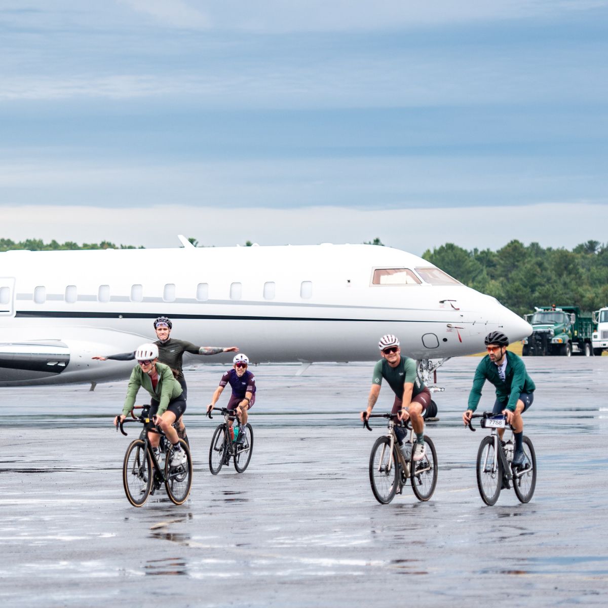 A group of cyclists rides on a wet, reflective runway. A white private jet is parked in the background. The sky is overcast, and the cyclists wear helmets and activewear.