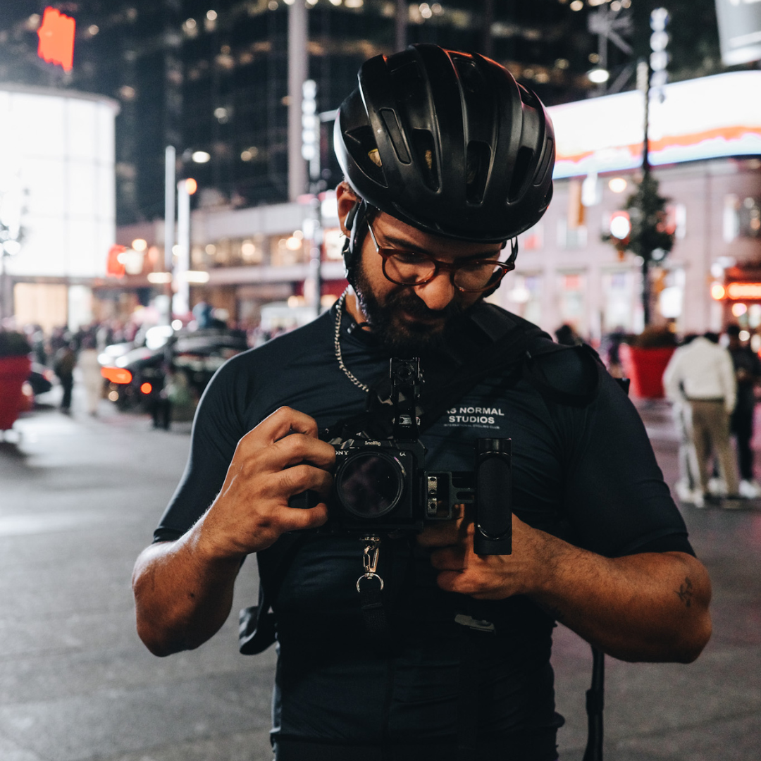 A man in a helmet, standing in a bustling city street at night, looks intently at a camera in his hands. The street is busy with people and lit by neon signs and building lights, creating a lively urban atmosphere.