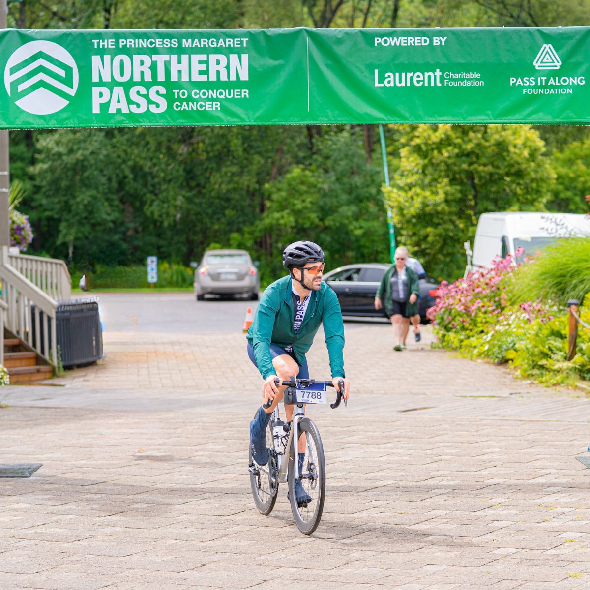 A cyclist wearing a helmet and green jacket rides under a banner that reads "The Princess Margaret Northern Pass to Conquer Cancer." The event is supported by Laurent Charitable Foundation and Pass It Along Foundation. Lush greenery surrounds the area.