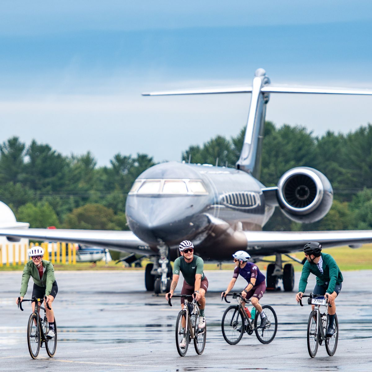 Four cyclists ride their bikes on an airfield in front of a large private jet. The sky is overcast, and trees are visible in the background.