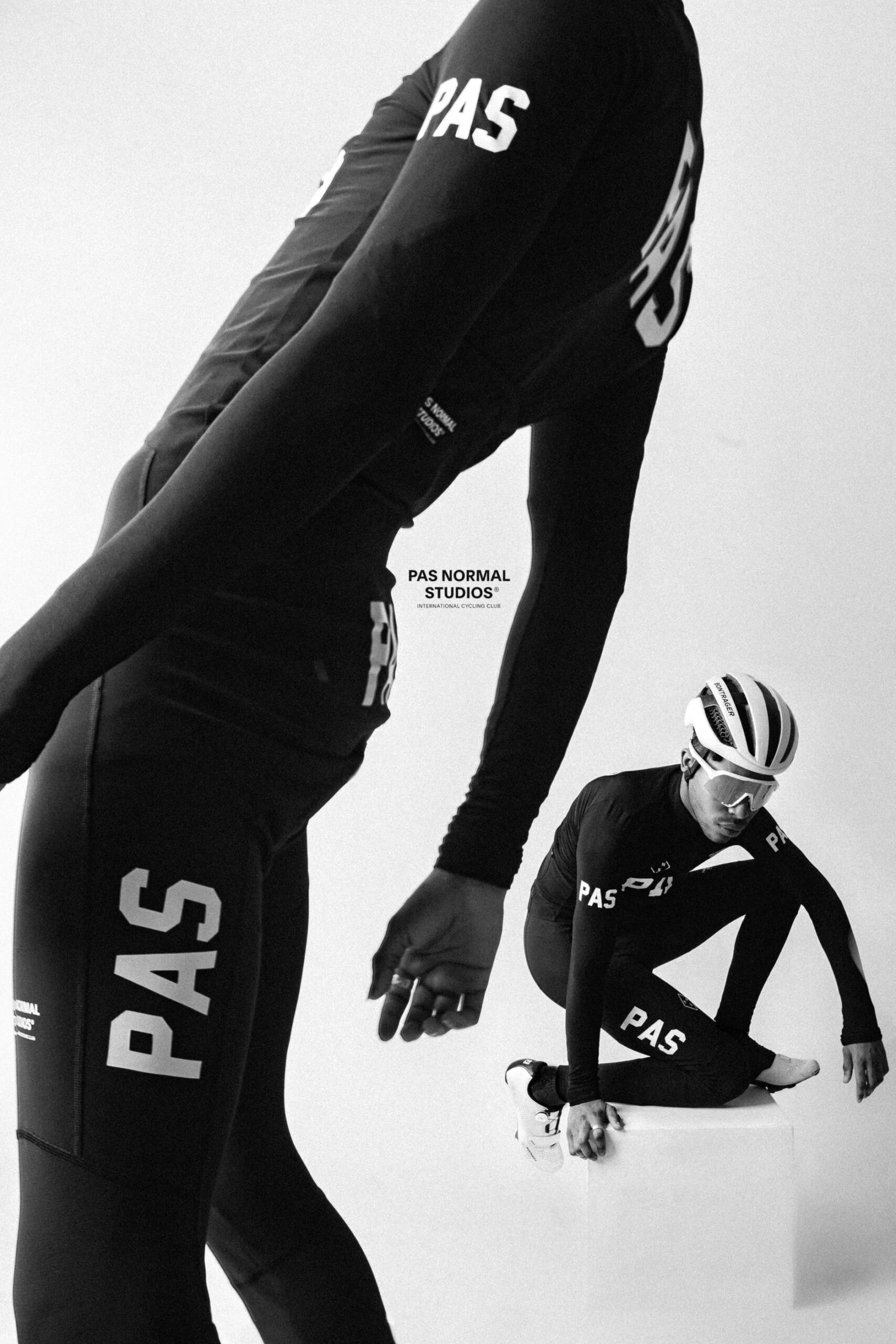 Two cyclists wearing PAS Normal Studios gear pose in black-and-white. One stands leaning forward, while the other sits on a block, wearing a helmet. Bold "PAS" logos are visible on their apparel.