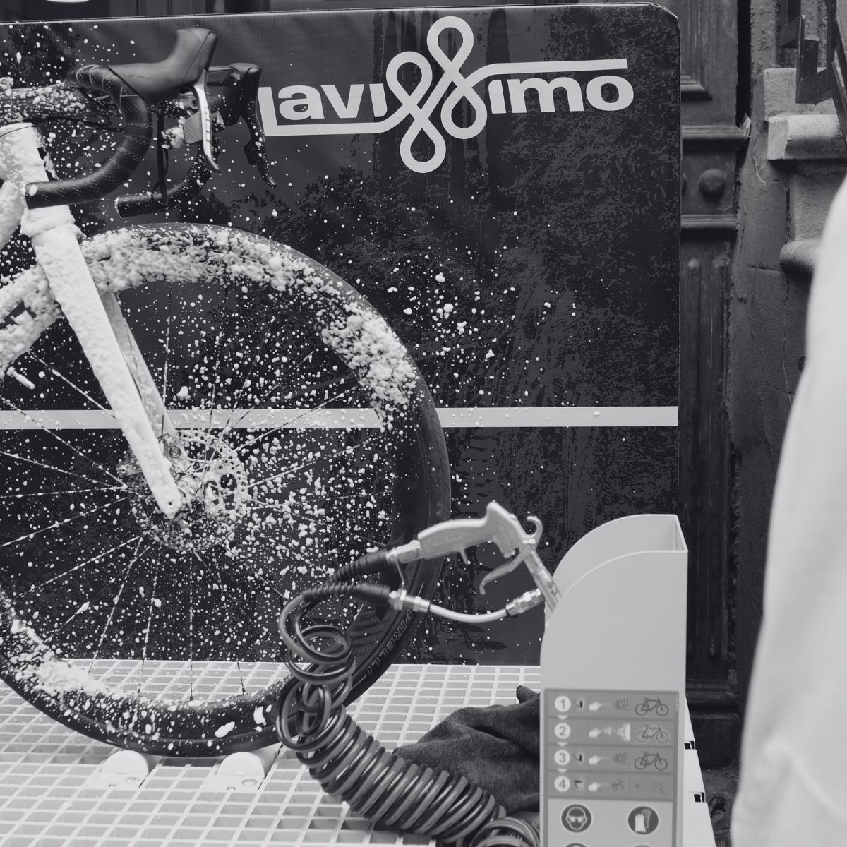 A bicycle covered in soap suds is mounted on a stand for cleaning. A washing station, labeled "lavàmio," displays a control panel with buttons for various settings. The scene is in black and white, focusing on the bike and cleaning equipment.