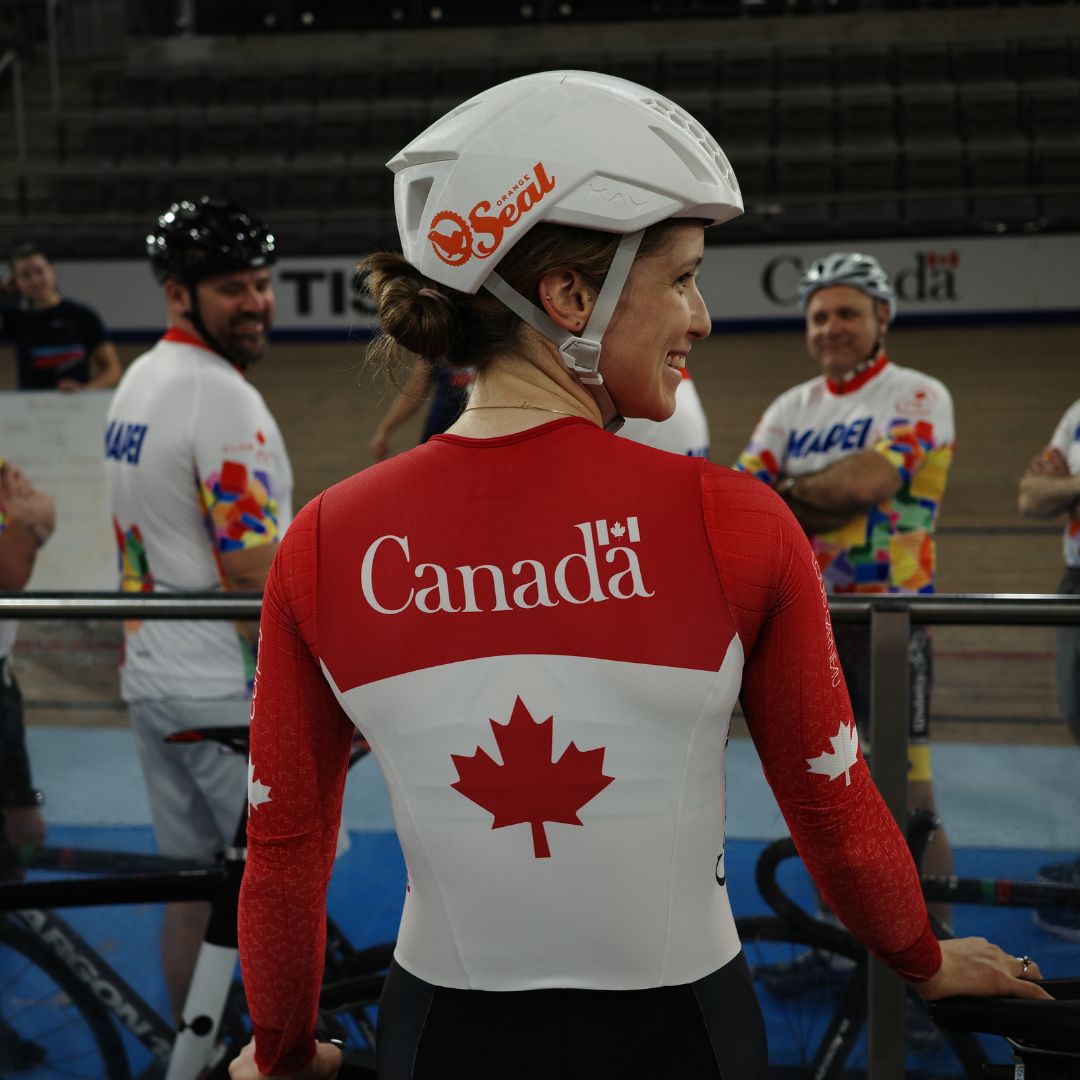 A cyclist in a red and white Canadian jersey with a maple leaf design stands smiling. She wears a white helmet and is surrounded by other cyclists and spectators in an indoor track cycling arena.