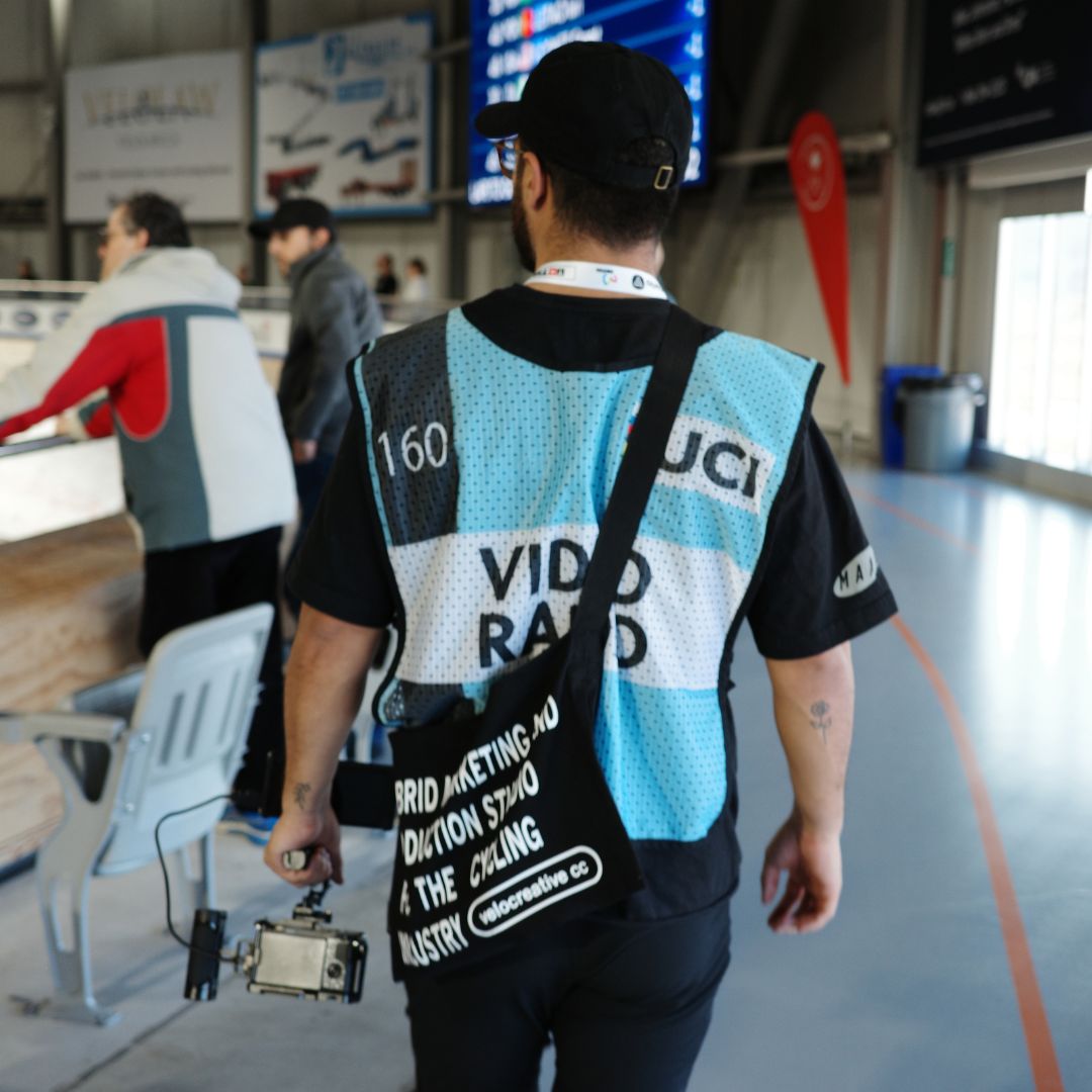 A person wearing a blue and white "UCI VIDEO RADIO" vest walks indoors on a track, carrying a camera. Other people and indoor signage are visible in the background.