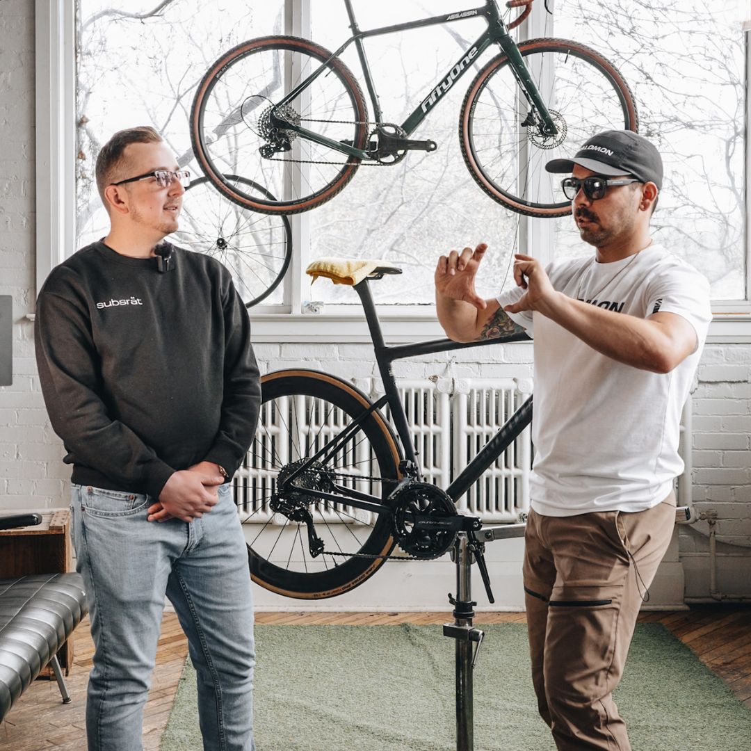 Two people are in a room with a bicycle mounted on a stand and another hanging on the wall. One person is explaining something while gesturing with his hands, and the other is listening attentively. The room has a window and a sofa.