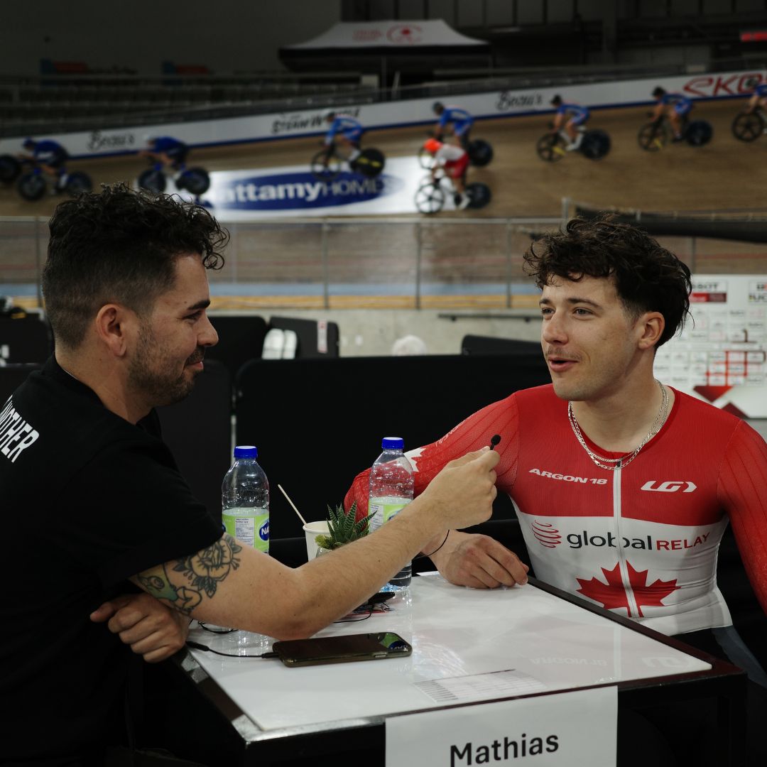 A cyclist in a red and white jersey with "Global Relay" sits at a table labeled "Mathias" being interviewed by a man holding a microphone. Cyclists speed around the indoor track in the background.