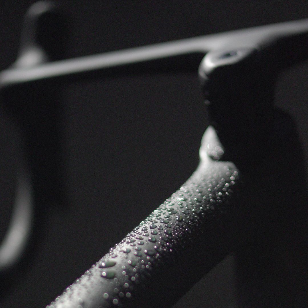 Close-up of a bicycle frame with water droplets on a dark, matte surface. The lighting highlights the droplets, creating a contrast against the dark background. The image captures the texture and sleek design of the bike frame.