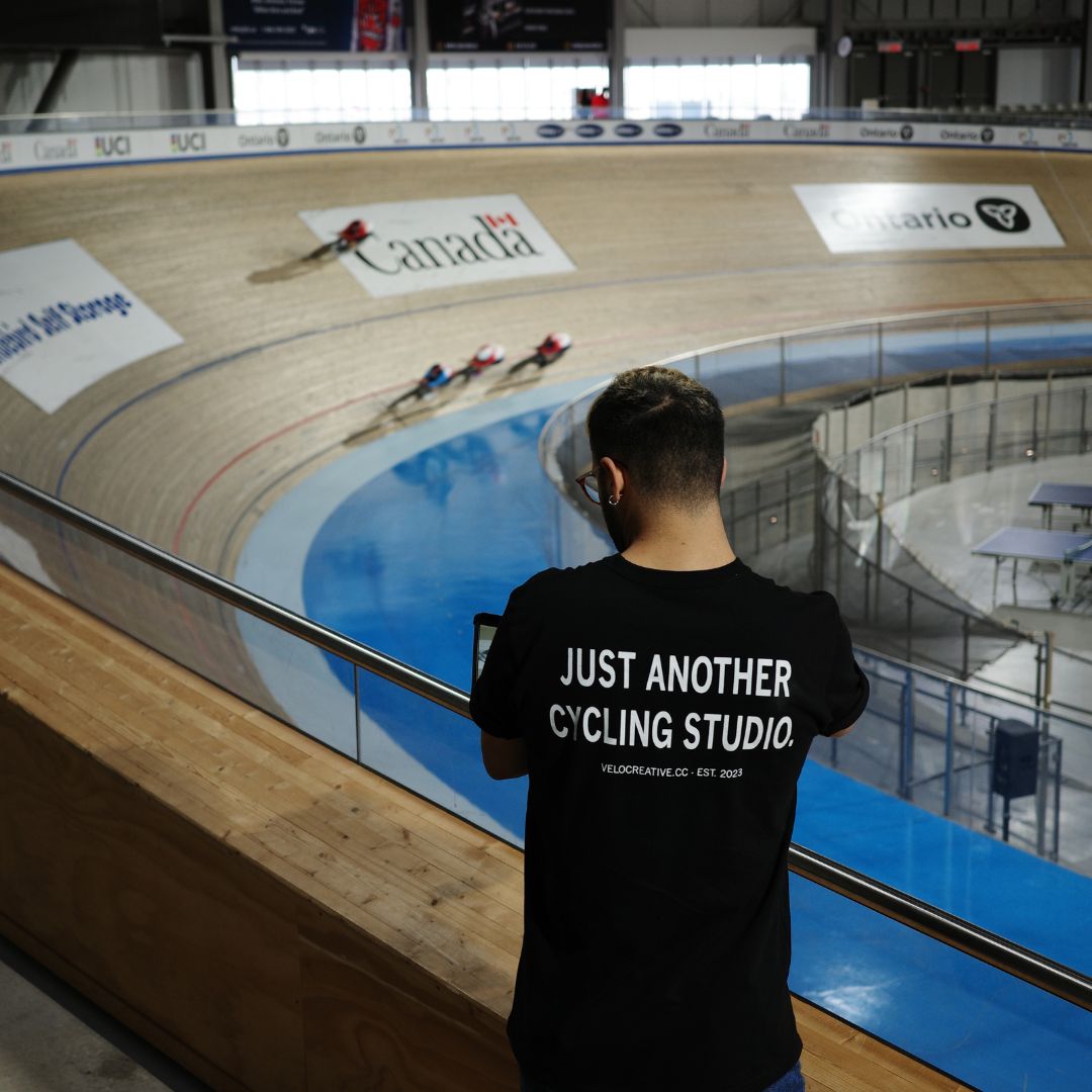 A person wearing a shirt with the text "Just Another Cycling Studio" stands overlooking a velodrome track. Cyclists in red uniforms race on the curved wooden track. The venue has banners and seating areas in the background.