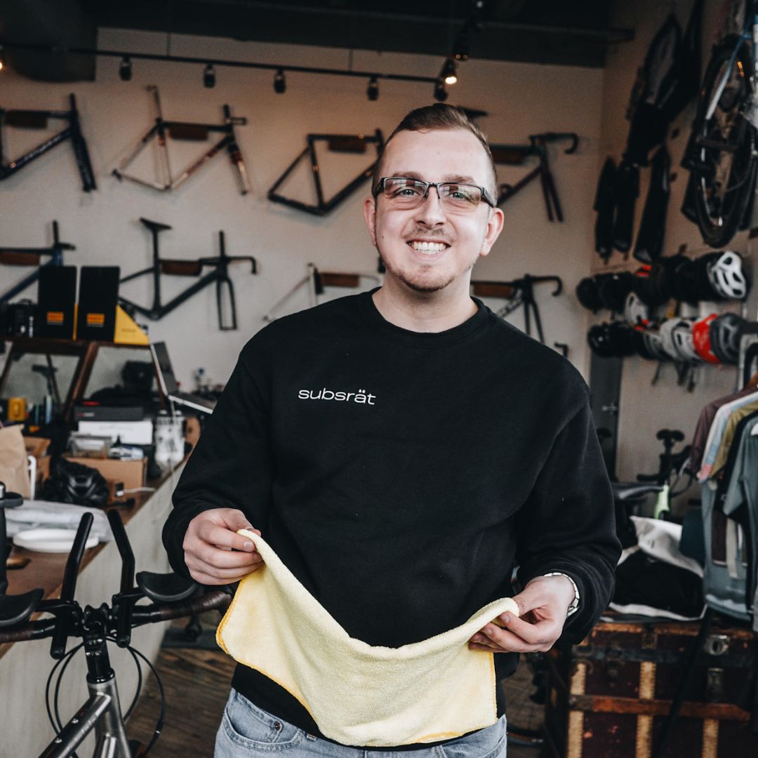 A person wearing glasses and a black sweatshirt with "subsät" on it holds a yellow cloth, smiling inside a bicycle shop. The background features bicycle frames and helmets displayed on the walls.