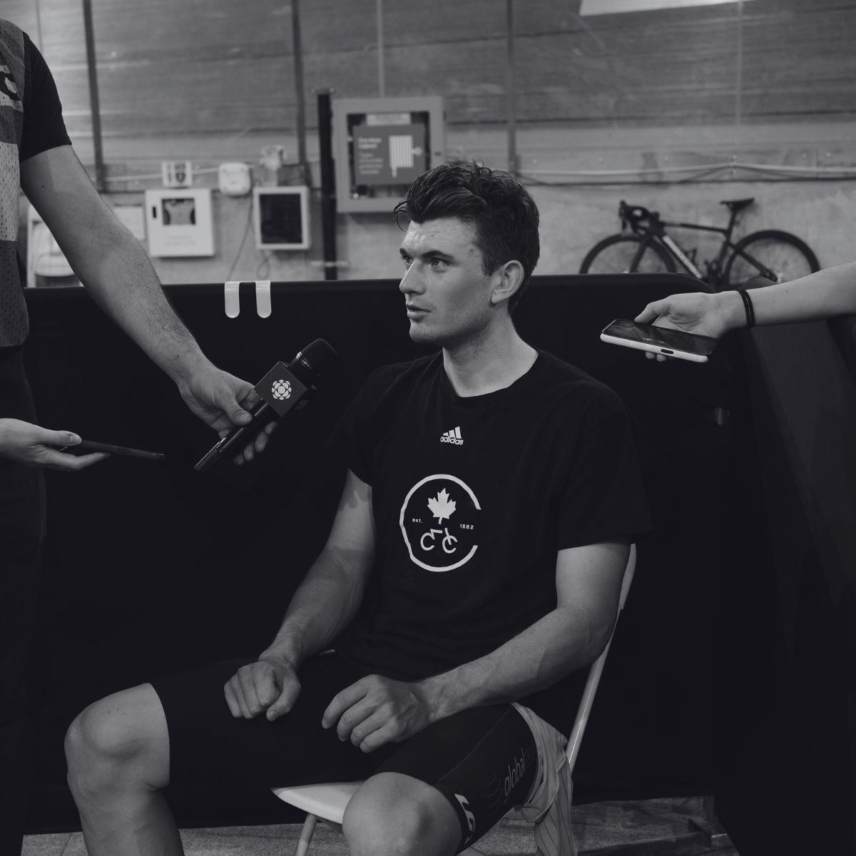 A seated athlete wearing a black t-shirt with a maple leaf logo is being interviewed by two reporters holding microphones and smartphones. A bicycle is visible in the background.