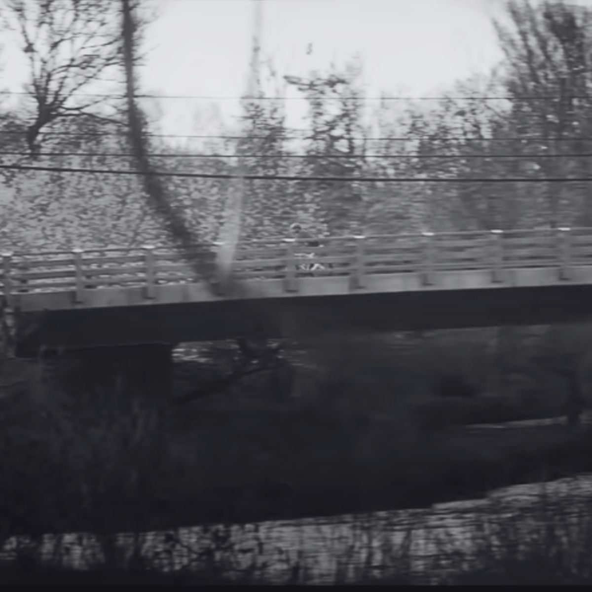 A black and white image of a bridge crossing over a stream. Trees with bare branches fill the background, indicating a winter or early spring setting. The bridge is empty, creating a serene and quiet atmosphere.