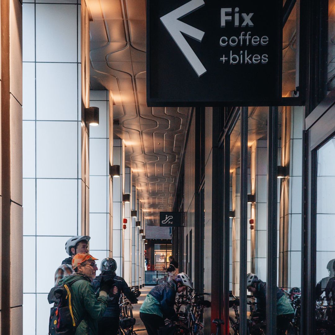 People with bicycles stand outside a shop with a sign reading "Fix coffee + bikes." The shop has a modern design with white paneling and a patterned ceiling.