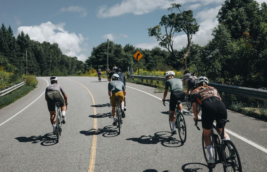 A group of cyclists riding on a scenic, curving road surrounded by trees and greenery under a partly cloudy sky. The cyclists wear helmets and are spaced out along the road, with a few more bikers visible in the distance.