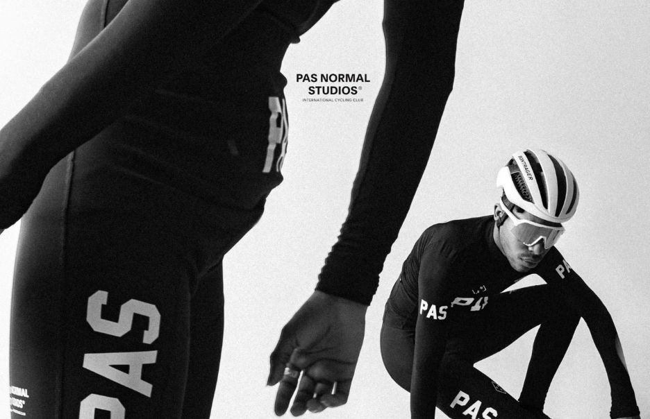 Black and white image of two cyclists wearing PAS Normal Studios cycling gear. One cyclist is in the foreground showing part of their leg, while the other is crouched with a helmet and glasses, looking to the side. The brand logo is visible in the background.