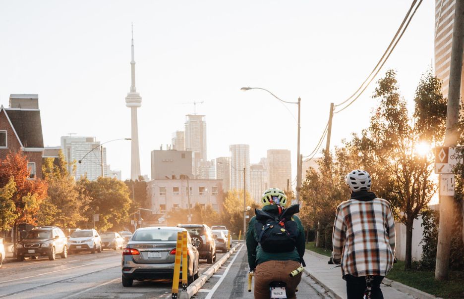 Two cyclists ride on a city street lined with traffic and buildings. The skyline features a prominent tower in the background, and the sun casts a warm glow over the scene.