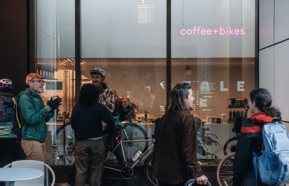 A group of people in casual clothing stand outside a shop with a glass window, through which bicycles and a neon sign reading "coffee+bikes" are visible. Some people are wearing helmets and carrying backpacks.