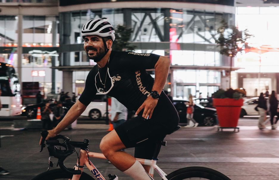 A cyclist in a helmet and black outfit is riding a bike through a busy city street at night. The background features a modern building with bright lights and people walking.