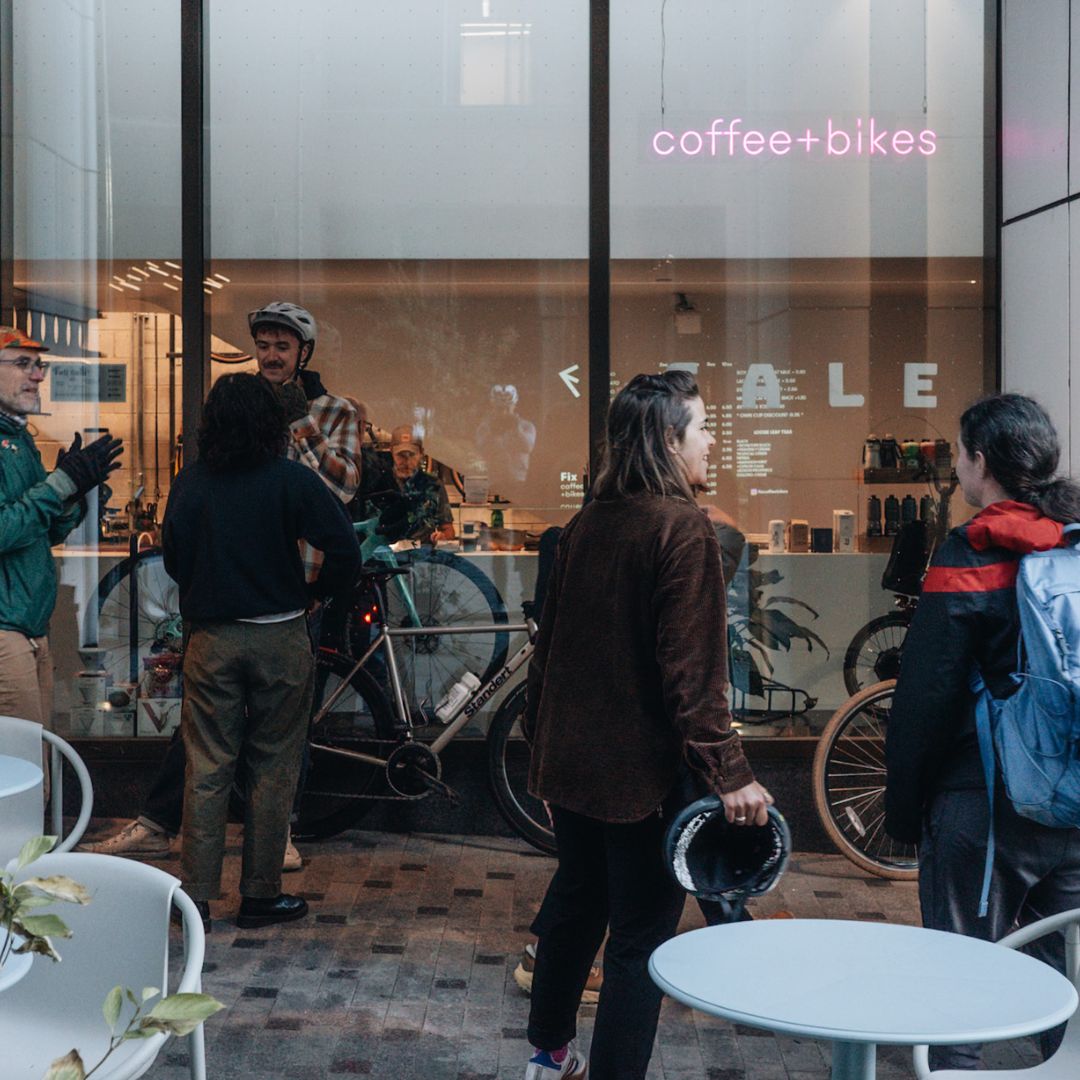 A group of people stand outside a coffee shop named "coffee+bikes." Some are holding helmets and a bicycle is visible. They appear to be in conversation. Tables and chairs are arranged around the entrance.