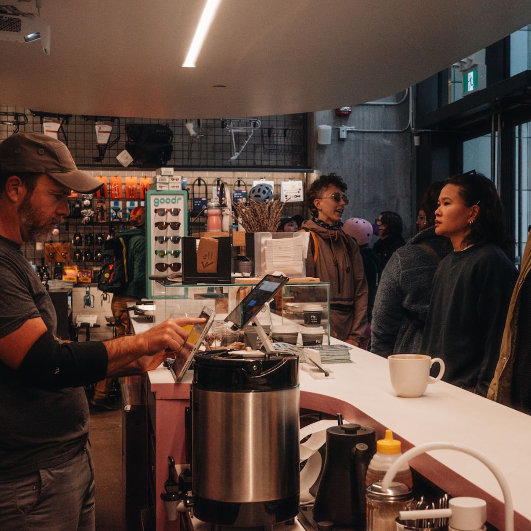 A barista wearing a cap and arm brace takes an order on a tablet at a cozy cafe. Customers line up beside the counter, awaiting service. The cafe is filled with merchandise and has a warm, inviting atmosphere.