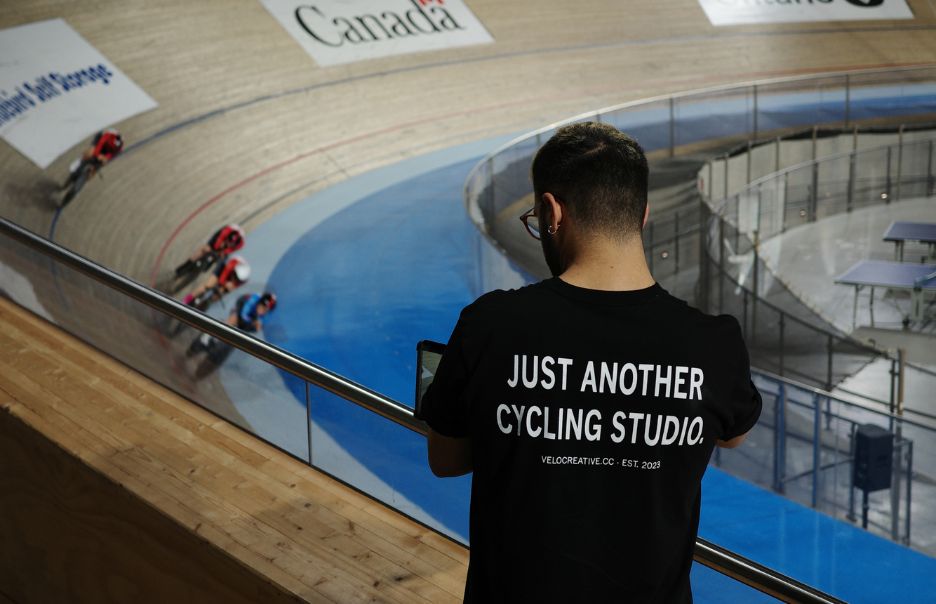 A man wearing a black "Just Another Cycling Studio" t-shirt watches a cycling race at an indoor velodrome. Several cyclists in red gear race on the curved track. The word "Canada" is visible on a banner in the background.