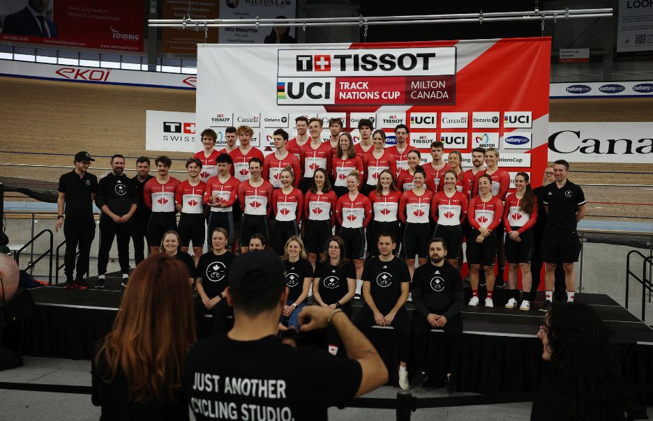 A large group of athletes in red and white uniforms, likely representing Canada, pose in front of a banner for the TISSOT UCI Track Nations Cup in Milton, Canada. Coaches and staff in black outfits are seated in front.