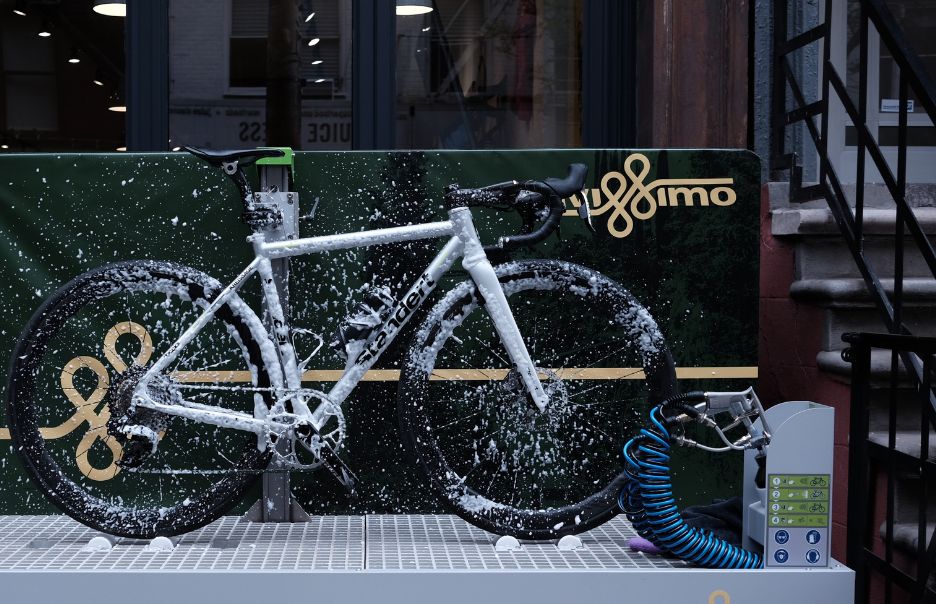 A bicycle covered in soapy foam is placed on a wash platform outside a building. The background features large windows and a staircase. The bike is being cleaned, indicated by various cleaning equipment and hoses nearby.