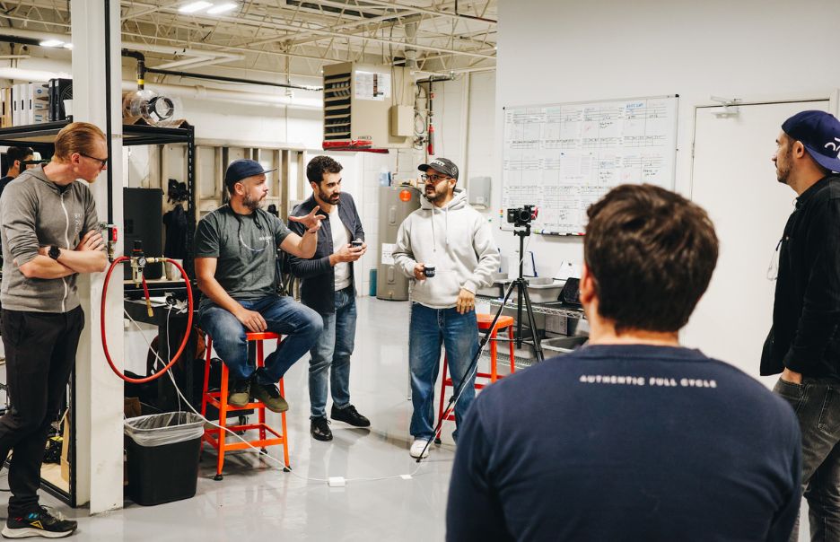 A group of six people are gathered in a workshop setting. Some are seated on stools, and a tripod-mounted camera is nearby. They appear to be having a discussion, surrounded by industrial equipment and a whiteboard.