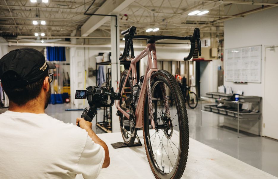 A person using a camera to film a bicycle on a stand in a workshop. The bicycle is a gravel bike with drop bars and knobby tires. The workshop has tools and equipment visible in the background.