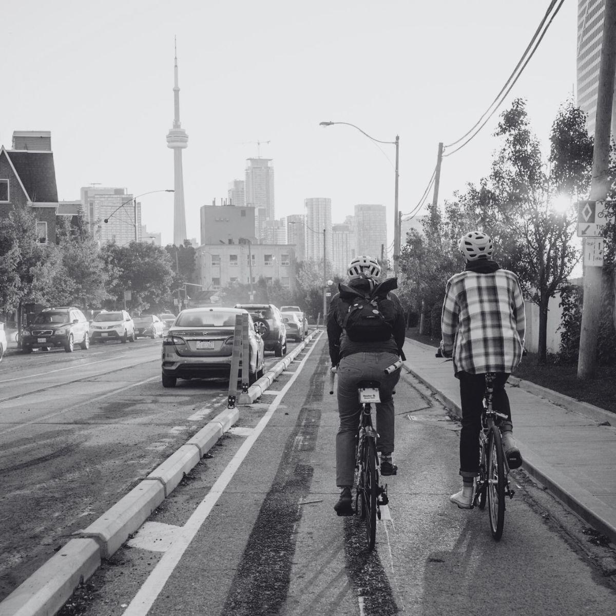 Two cyclists ride along a city street in the bike lane, with cars on the left. The skyline, featuring the CN Tower, is visible in the background. The scene is in black and white.