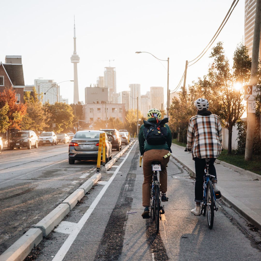 Two cyclists ride on a city street bike lane. The skyline, including a prominent tower, is visible in the background against a setting sun. Cars are on the adjacent road, and trees line the sidewalk.