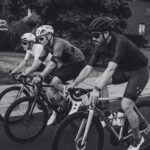 Three men ride bicycles on a street, wearing cycling attire and helmets. They are focused and moving in close formation. The scene is in black and white, with trees and a building visible in the background.