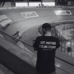 A man wearing a shirt that says "Just Another Cycling Studio" watches from the sidelines of an indoor velodrome as cyclists race on the track. The curved wooden track and various logos are visible in the background.