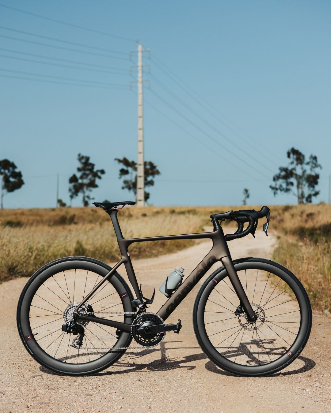 A sleek, modern road bike with a water bottle rests on a gravel path. The background features dry grass, sparse trees, a tall utility pole, and a clear blue sky.