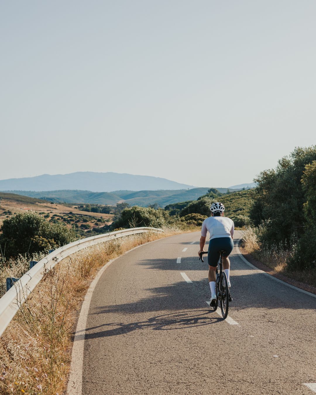 A cyclist wearing a white helmet and shirt rides along a winding road through hilly, green landscape under a clear sky.