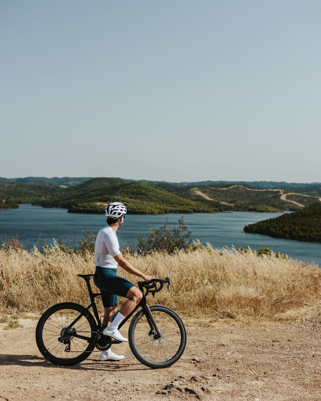 A cyclist wearing a helmet and sunglasses stops on a dirt path overlooking a scenic lake surrounded by hills and dry grass under a clear blue sky.