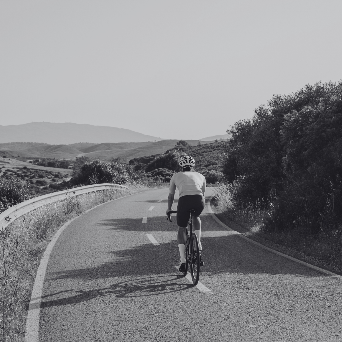 A cyclist wearing a helmet rides alone on a winding road through a hilly landscape with trees and distant mountains, under a clear sky. The image is in black and white.