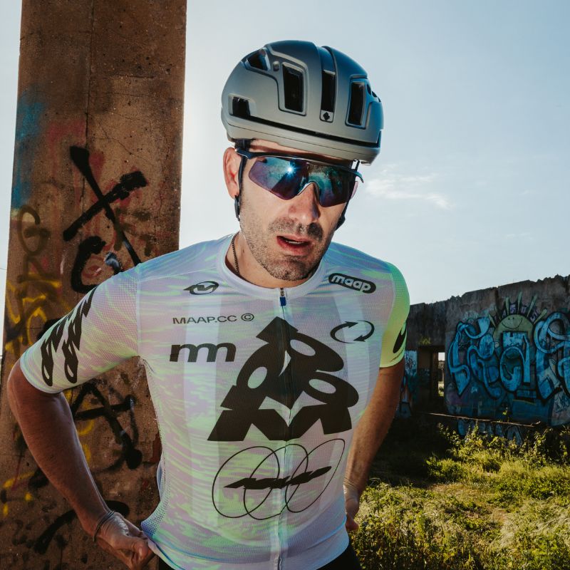 A cyclist in a helmet, sunglasses, and a patterned jersey stands outdoors by a graffitied pillar. Sunlight highlights his face, and there is graffiti and grass in the background.