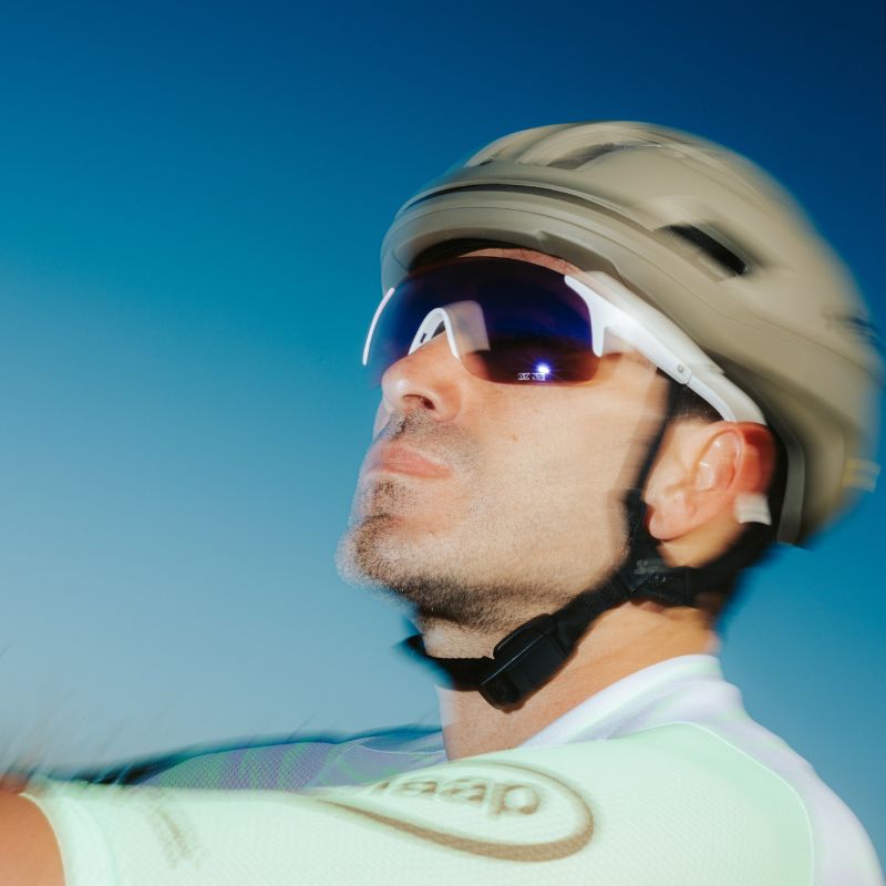 A man wearing a helmet, sunglasses, and a light cycling jersey looks ahead outdoors, with a blurred blue sky background.