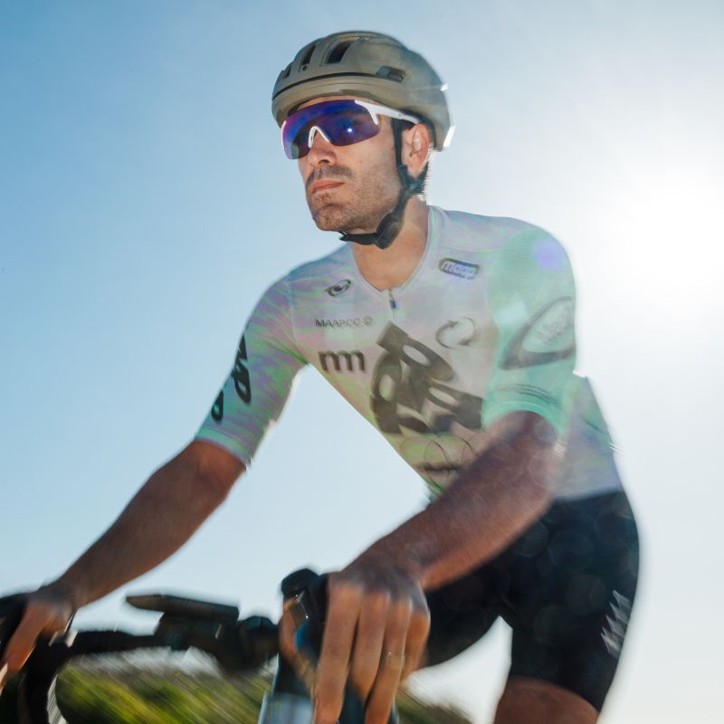 A cyclist wearing a helmet, sunglasses, and a light-colored jersey rides a bike outdoors on a sunny day, with the sun shining brightly behind him.